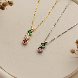 May include: Two delicate necklaces with pendant designs. One is gold-toned, the other silver-toned. Each pendant features three square-cut gemstones in a vertical row. The gold necklace has green, white, and red stones. The silver necklace has green, pink, and green stones.