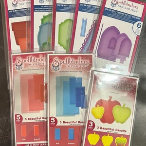 May include: A collection of Spellbinders Nestabilities die-cut systems for crafting. The dies are packaged in clear plastic sleeves with colourful images of the shapes they create. The dies include a variety of shapes, such as rectangles, arches, apples, and labels. The packaging also includes the text "Use in most leading die-cut systems."