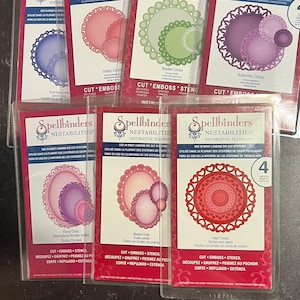 Puede incluir: Un conjunto de seis elementos de troquelado decorativos Spellbinders Nestabilities para manualidades. Los troqueles están empaquetados en fundas de plástico transparente con etiquetas rosas y blancas. Las etiquetas incluyen el logotipo de Spellbinders, el nombre del producto y las instrucciones de uso. Los troqueles están diseñados para crear formas intrincadas, como círculos, óvalos y mariposas.