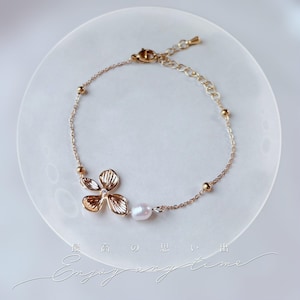 Peut inclure: Bracelet doré orné d'une fleur avec une petite pierre précieuse transparente au centre. Il comprend une perle d'eau douce et de petites perles dorées. Les mots "Enjoy any time" sont écrits en cursive.