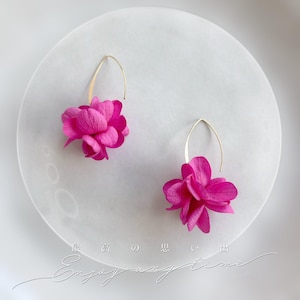 Può includere: Un paio di orecchini floreali rosa acceso con ganci color oro. Gli orecchini sono composti da petali raggruppati e stratificati, creando un aspetto pieno e strutturato. Sono presentati su una superficie circolare bianca.