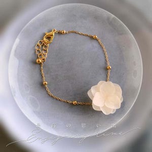 May include: A gold chain bracelet featuring a delicate white flower charm. The bracelet has a lobster clasp fastening and small round beads along the chain. The flower charm is made of layered petals and has a soft, feminine look.