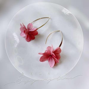 Könnte beinhalten: Ein Paar goldfarbene Creolen mit Clustern aus konservierten rosa und roten Hortensienblütenblättern. Die Ohrringe sind auf einer durchscheinenden kreisförmigen Oberfläche mit dem Text "最高の思い出" und "Enjoy any time" abgebildet.