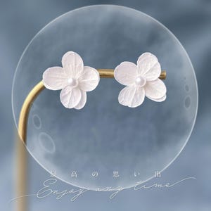Op de afbeelding: Een paar delicate witte bloemvormige oorknopjes met een parel in het midden, gezet op een goudkleurige staaf. De oorbellen worden gepresenteerd tegen een zachte blauwe achtergrond, met de woorden "Enjoy any time" in cursief.
