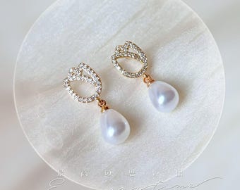 May include: A pair of gold-toned earrings featuring a tulip-shaped design encrusted with small, clear crystals. Each earring has a teardrop-shaped, white pearl pendant. The earrings are displayed on a white, textured surface.