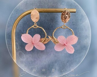 Può includere: Orecchini dorati con un delicato design circolare e un fiore rosa pressato. Gli orecchini sono esposti su un supporto dorato.