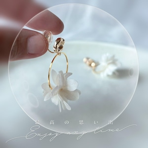 Può includere: Orecchini a cerchio color oro con un delicato design a fiore bianco. Gli orecchini sono tenuti da una mano, mostrando gli intricati dettagli della composizione floreale. Gli orecchini sono su una superficie bianca. Il testo "Enjoy any time" è visibile.