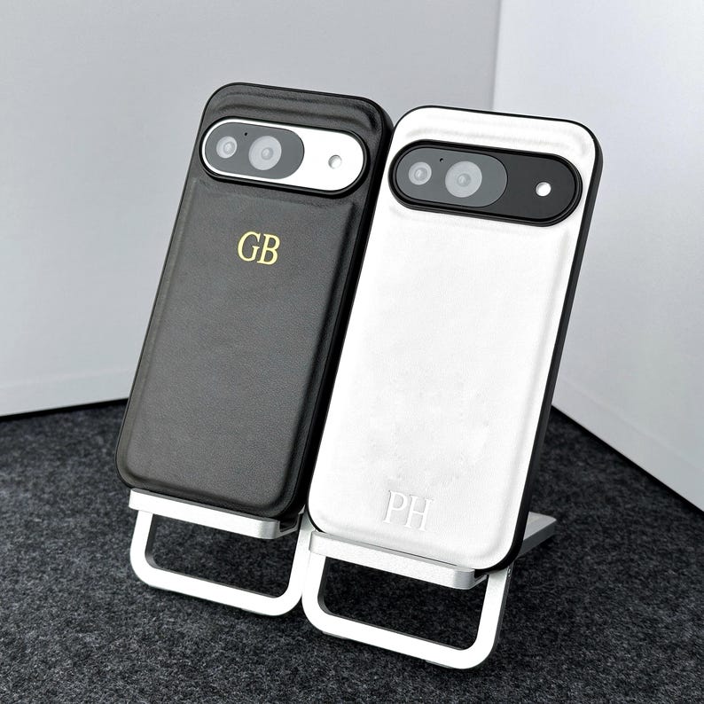 May include: Two black leather phone cases with a silver stand. The case on the left has the initials "GB" embossed on the back. The case on the right has the initials "PH" embossed on the back.