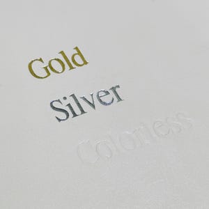 May include: Close-up of a white surface with the words "Gold", "Silver", and "Colorless" embossed in different colours.