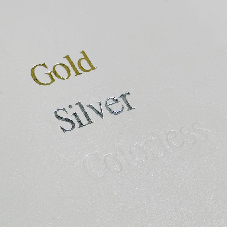 May include: Close-up of a white surface with the words "Gold", "Silver", and "Colorless" embossed in different colours.