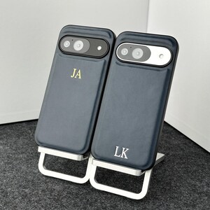 May include: Two dark blue phone cases with initials "JA" and "LK" in gold lettering. The cases are on a silver stand.