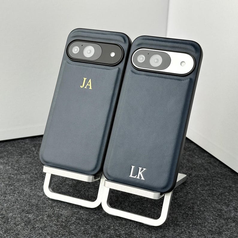 May include: Two dark blue phone cases with initials "JA" and "LK" in gold lettering. The cases are on a silver stand.