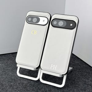 May include: Two white phone cases with black trim and a silver stand. The cases have the initials "CB" and "PH" engraved on them.