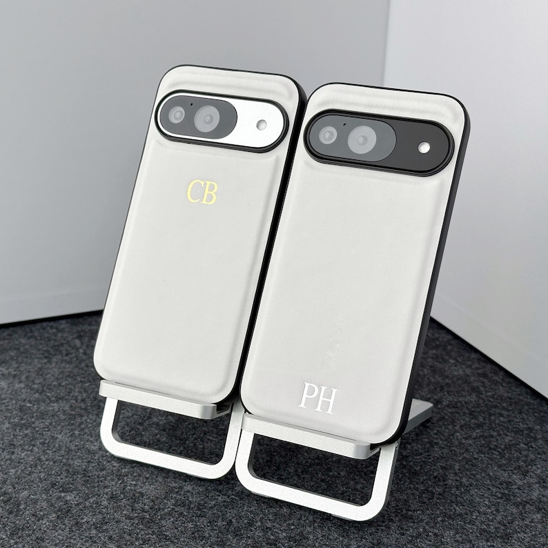 May include: Two white phone cases with black trim and a silver stand. The cases have the initials "CB" and "PH" engraved on them.