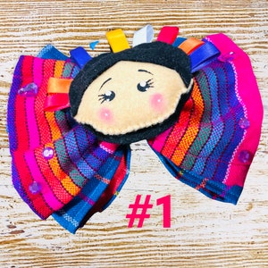 May include: A colorful handmade fabric bow with a felt doll face in the center. The bow is made of a striped fabric with pink, blue, yellow, and orange stripes. The doll face has black hair, brown eyes, and pink cheeks. The bow is decorated with purple and clear beads. The number "#1" is written in pink below the bow.