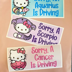 May include: Three Hello Kitty car stickers with different zodiac signs. The first sticker is blue and says "Sorry, An Aquarius Is Driving". The second sticker is purple and says "Sorry, A Scorpio Is Driving". The third sticker is pink and says "Sorry, A Cancer Is Driving".