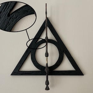May include: A black wooden Deathly Hallows symbol with a wand. The symbol is a triangle with a circle in the middle and a vertical line. The wand is dark brown with a detailed handle.