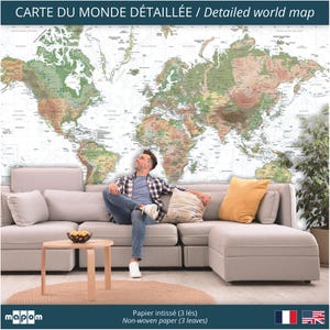 Pode incluir: Um mural de papel de parede com um mapa mundial detalhado em um ambiente de sala de estar. O mapa &eacute; impresso em papel n&atilde;o tecido e est&aacute; dispon&iacute;vel em tr&ecirc;s pain&eacute;is. O mapa apresenta uma combina&ccedil;&atilde;o de cores marrom claro e verde sobre um fundo branco. Uma pessoa est&aacute; sentada em um sof&aacute; seccional cinza claro em frente ao mapa.