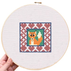 May include: A cross-stitch embroidery hoop with a red fox design. The fox is surrounded by a red, white, and blue patterned border.