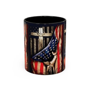 May include: Black ceramic mug featuring an American flag design with a hand holding the flag. The design includes a cross and stars and stripes. The mug has a black interior and a white background.