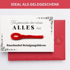 May include: A white card with the text "Für jemanden der schon ALLES hat" and "Bauchnabel Reinigungsbürste" with a red pipe cleaner in the center. The card is placed on top of a red envelope.