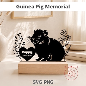 May include: A black guinea pig memorial with the text "Guinea Pig Memorial" at the top. The memorial features a silhouette of a guinea pig, a heart with the name "Poppy" and dates, and floral accents. It sits on a wooden base.