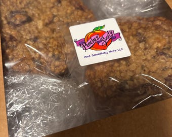 May include: Two round, textured oatmeal cookies packaged in a brown cardboard box with a clear plastic window. A square white sticker on the plastic features a peach graphic with the text Peaches Sweets And Something More LLC in purple.