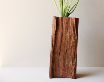 Puede incluir: Un jarrón de madera con una textura rústica, similar a la corteza, contiene un ramo de flores rosas y naranjas con tallos verdes.