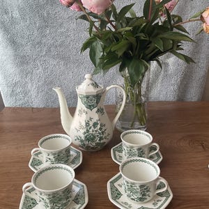 May include: A white porcelain tea set featuring a green floral pattern, consisting of a teapot and four matching cups with saucers. The teapot has an octagonal shape, and each cup and saucer set displays intricate botanical illustrations.