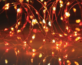 Puede incluir: Luces de cadena LED Sunset de Qbis UK. Primer plano de una cadena de luces de alambre de cobre con pequeñas bombillas LED de tonos cálidos. Las luces son una mezcla de naranja y amarillo, creando un brillo cálido. El texto "Sunset LED String Lights by Qbis UK" es visible.
