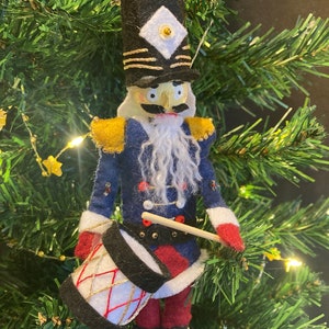 May include: A felt nutcracker ornament with a blue uniform, gold trim, and a white beard. He is holding a drum and a drumstick.