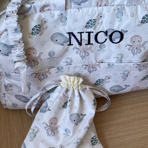 May include: White canvas travel bag with a drawstring closure and a zippered top. The bag has a white and blue print of sea creatures, including whales, octopuses, and seahorses. The bag is personalized with the name "NICO" in black embroidery.