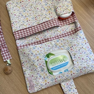 May include: A fabric pouch featuring a floral pattern with small colorful flowers on a white background, accented by a pink and white gingham trim. The pouch includes a central opening for a wet wipe pack labeled Papel WC Higiénico Húmedo.