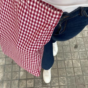 May include: A red and white gingham patterned tote bag made of fabric, featuring a classic checkered design. The bag has two shoulder straps and a rectangular shape, shown in a top-down view against a background of blue denim jeans and white sneakers.