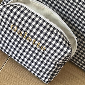 Puede incluir: Un neceser con estampado de cuadros vichy en blanco y negro con cremallera blanca y la palabra "Galette" bordada en dorado. La bolsa está abierta, revelando un interior blanco. Una bolsa a juego está en el fondo.