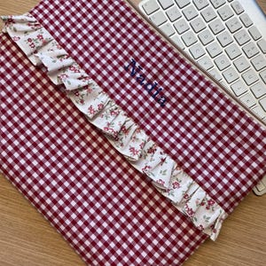 Könnte beinhalten: Eine rechteckige Stofftasche mit rot-weißem Vichy-Muster und Reißverschluss. Eine Rüschenborte mit kleinem Blumenmuster verläuft diagonal über die Vorderseite. Der Name Nadia ist mit dunkelblauem Garn auf den Stoff gestickt.