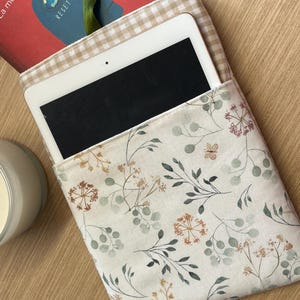 Puede incluir: Una funda de tableta color crema con un estampado floral con hojas verdes y flores marrones. La funda está abierta, revelando una tableta blanca. Un libro con la palabra "RESET" es parcialmente visible.