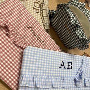 Puede incluir: Tres estuches rectangulares con cremallera y un bolso pequeño redondeado con estampado de cuadros vichy en beige, rojo, azul y verde. Cada artículo está adornado con un borde de tela con volantes y personalizado con texto bordado como CCP, Français y AE.
