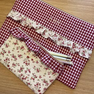 Puede incluir: Un estuche rectangular grande con patrón de cuadros vichy rojo y blanco y un estuche rectangular más pequeño con estampado floral crema. Ambos tienen un volante, cremallera dorada y las letras ABV bordadas en la parte frontal.