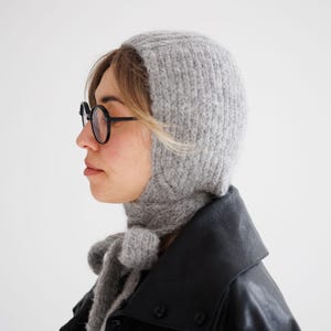 May include: A light grey knitted balaclava hood with a textured, fuzzy surface. The headpiece features long, integrated knitted ties that hang down from the chin area, allowing for a secure fit under the jawline with a simple knot.