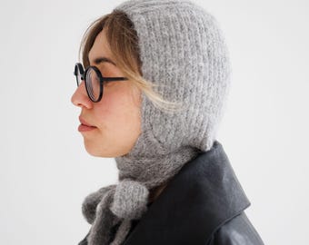 May include: A light gray knitted balaclava hood with a textured, fuzzy surface. The headpiece features long, integrated knitted ties that hang down from the chin area, allowing for a secure fit under the jawline with a simple knot.
