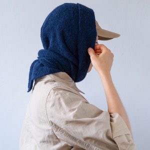 May include: A person wearing a blue knitted scarf over their head and a beige shirt. They are wearing a brown baseball cap.