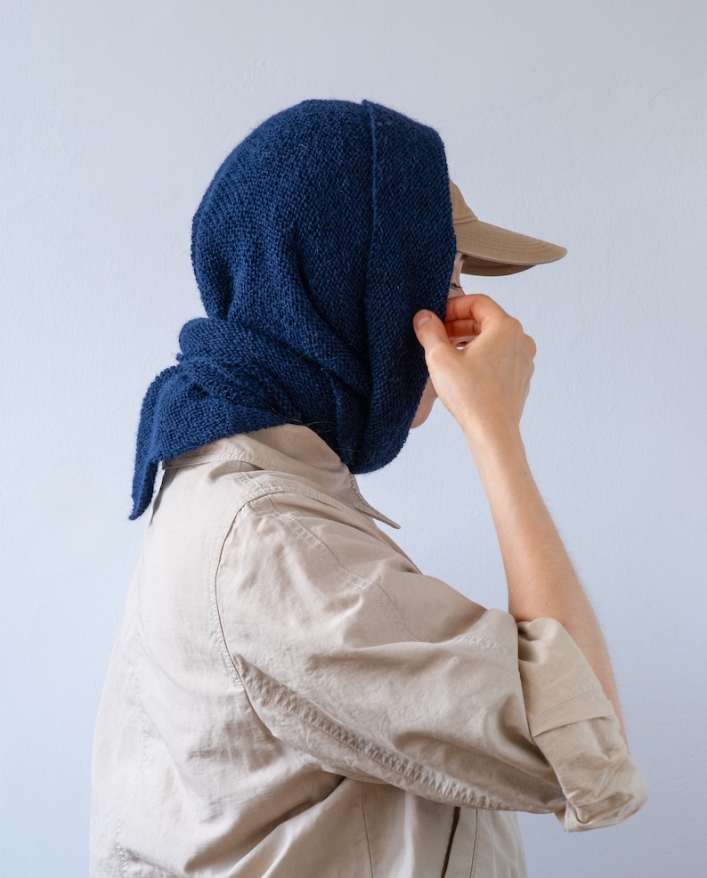 May include: A person wearing a blue knitted scarf over their head and a beige shirt. They are wearing a brown baseball cap.