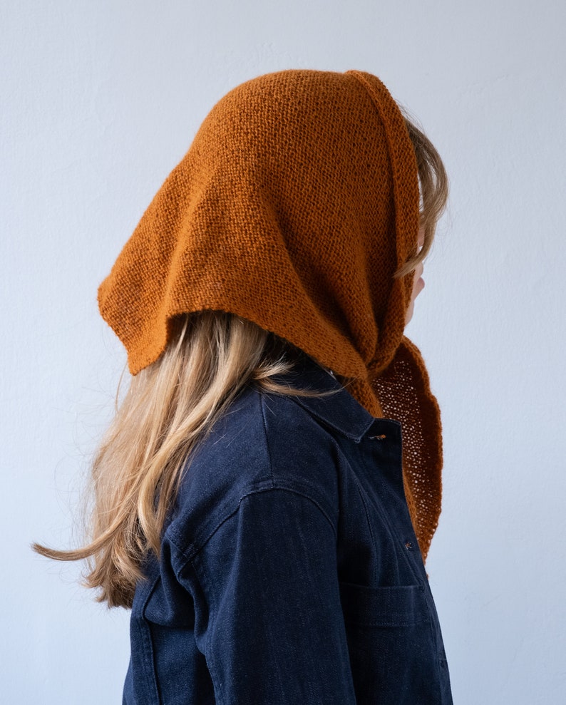 May include: A brown knitted scarf with a long, flowing end. The scarf is worn over the head and tied at the neck.