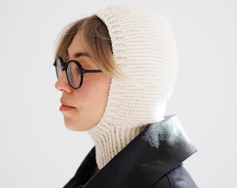 May include: A cream-colored knit balaclava featuring a ribbed texture around the neck and a smooth, tight-knit construction covering the head, worn with round black-rimmed glasses and a black leather jacket.
