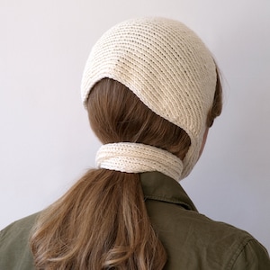 May include: A cream-colored knit bonnet with a ribbed texture and a long, integrated tie that wraps around the back of the head. The headwear features a rounded crown and side panels that cover the ears, crafted from a soft, textured yarn.