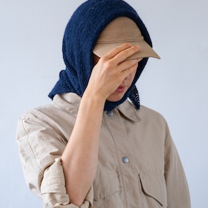 May include: A person wearing a beige shirt with a blue knitted scarf and a brown baseball cap.