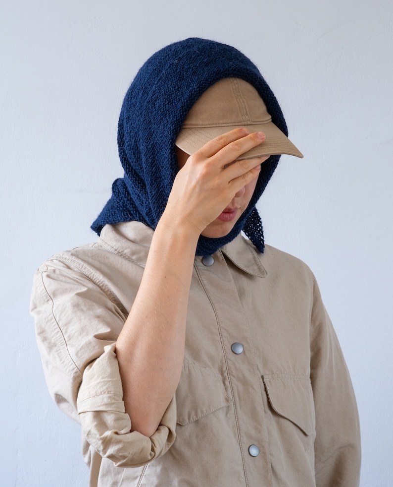 May include: A person wearing a beige shirt with a blue knitted scarf and a brown baseball cap.
