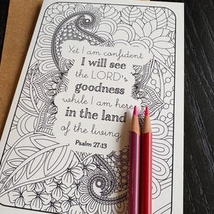 May include: A coloring card with a floral design and the text "Yet I am confident I will see the LORD's goodness...Psalm 27:13." Two colored pencils, one pink and one red, rest on the card. A brown envelope is in the background.