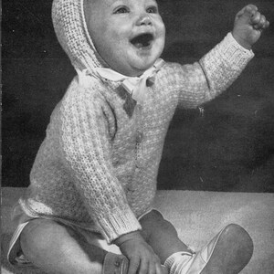 Peut inclure: Une photo en noir et blanc d'un bébé portant un pull en tricot blanc et un bonnet en tricot assorti. Le bébé est assis sur un tapis et sourit.